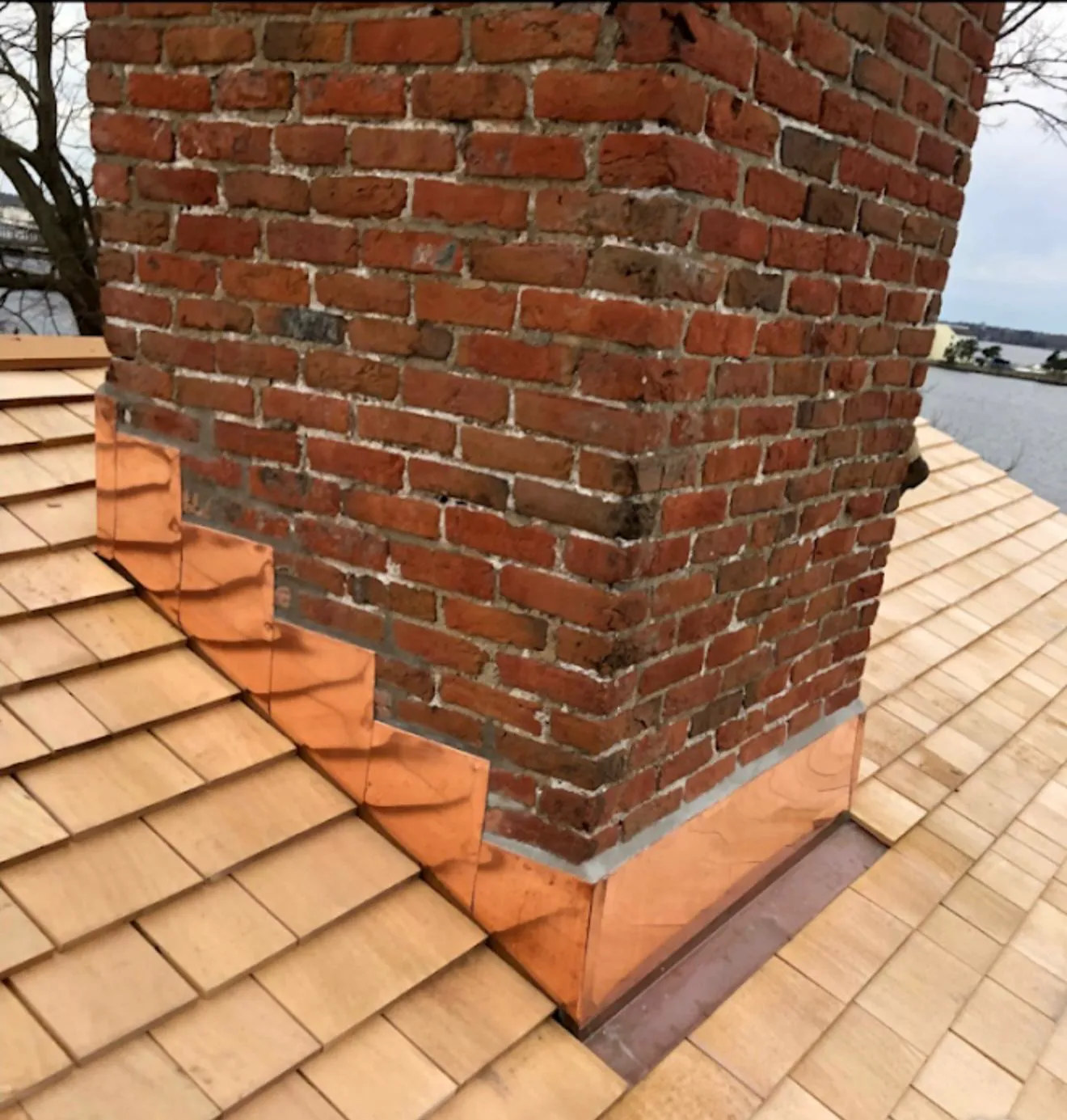 New copper step flashing sealing a brick chimney on a cedar-shake roof overlooking the water