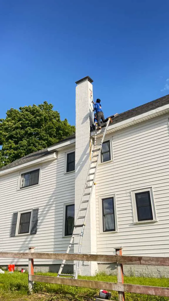 Technician on an extension ladder repairing a tall two-story painted brick chimney